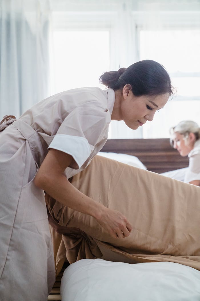 Hotel staff in uniform making a bed with precision and care in a bright room setting.